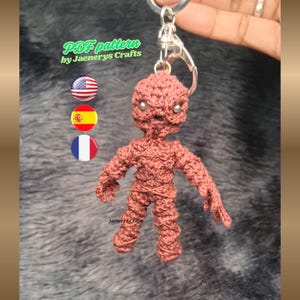 May include: A handmade, reddish-brown crochet doll keychain with silver-colored eyes and a silver keyring. The keychain is in the shape of a small figure with arms and legs. The text "PDF pattern by Jaenerys Crafts" is visible.