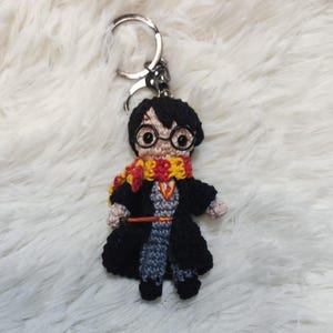 May include: A crocheted Harry Potter keyring with a black robe, red and yellow scarf, and round glasses. The keyring is attached to a silver key ring.