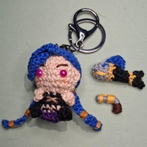 Puede incluir: Llavero de ganchillo con un personaje de pelo azul, ojos rosas y un atuendo negro. Incluye un pequeño objeto con forma de pez y una pistola en miniatura. El llavero tiene un cierre plateado.