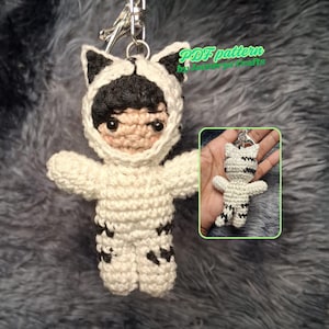 May include: A handmade crochet doll keychain in a white cat costume with black accents. The doll has black hair and is attached to a silver keychain. The image also includes a smaller version of the doll in a hand, with the text "PDF pattern by [seller's name] crafts".