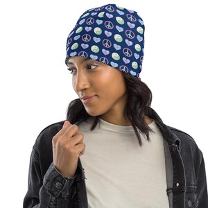 May include: A navy blue beanie featuring a repeating pattern of peace signs, hearts, and yin-yang symbols in pastel colors. The beanie is made of a soft material and is designed to be worn on the head.