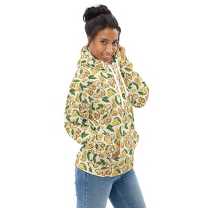 May include: A cream-colored hoodie with a repeating pattern of yellow ginger root and green leaves. The hoodie has a drawstring hood and a kangaroo pocket.