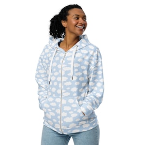 May include: A light blue zip-up hoodie with a white cloud pattern. The hoodie has a drawstring hood, a full-length zipper, and two front pockets. The model is wearing blue jeans.