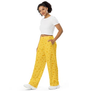 May include: Yellow cheese-patterned pants with a wide leg design. The pants have a repeating pattern of various sized holes, resembling Swiss cheese. The model is wearing a white crop top and white sneakers. The pants have pockets.