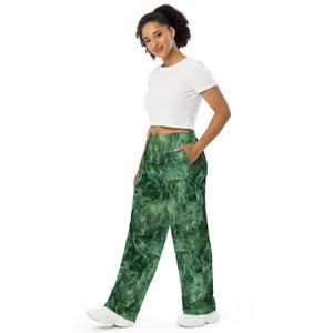 May include: Green patterned pants with a white top. The pants feature a swirling, smoke-like design in shades of green. The pants have pockets and a wide-leg cut. The model is wearing white sneakers.