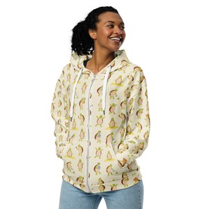 May include: A cream-colored zip-up hoodie with a repeating pattern of cartoon turtles holding paintbrushes. The turtles are green and brown with yellow accents. The hoodie has a hood and a zipper closure.