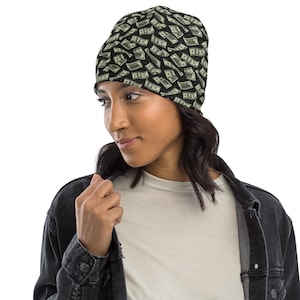 May include: A black beanie hat with a repeating pattern of US one-hundred dollar bills. The hat is worn by a person wearing a black denim jacket and a white t-shirt.