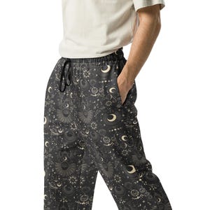 May include: Black pajama pants with a celestial pattern featuring moons, suns, and stars. The pants have a drawstring waist and pockets. The fabric is a dark gray with a repeating cream-colored design.