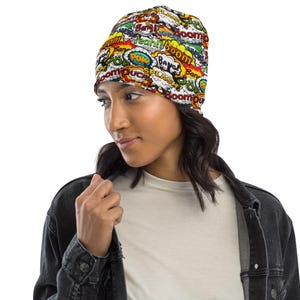May include: A colorful beanie featuring comic book sound effects like "Boom," "Pow," and "Bang!" in various speech bubbles. The beanie is predominantly white, red, yellow, and green. The wearer is wearing a black denim jacket.