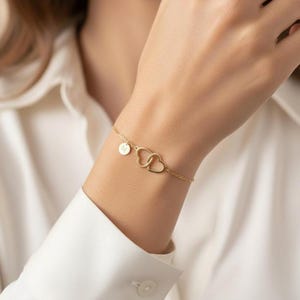 Op de afbeelding: Een delicate gouden armband met twee in elkaar grijpende hartvormige hangers en een kleine cirkelvormige bedel. De armband wordt om een pols gedragen, tegen een wit overhemd. De sieraden zijn eenvoudig en elegant.