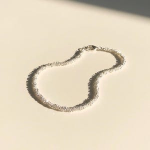 Può includere: Un braccialetto d'argento con un design strutturato e perlato. Il braccialetto è curvo e si trova su una superficie di colore chiaro, proiettando un'ombra. La chiusura è visibile a un'estremità.