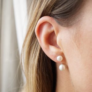 May include: A double-sided earring featuring two white spherical pearls of different sizes. The smaller pearl sits directly on the earlobe, while the larger pearl is positioned below the lobe, creating a layered effect with a minimalist aesthetic.