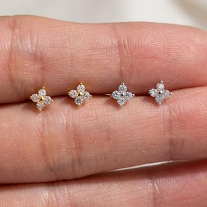 Può includere: Quattro piccoli orecchini a lobo disposti in fila, ognuno con un design a forma di fiore composto da cinque pietre rotonde trasparenti. I due orecchini a sinistra sono in metallo dorato, mentre i due a destra sono in metallo argentato.