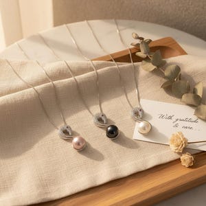 Può includere: Quattro collane d'argento con ciondoli dal design a forma curva ornati da piccole pietre trasparenti e una singola perla colorata. Le perle sono rosa, nere e bianche. È presente anche una carta con il testo "With gratitude & care..."