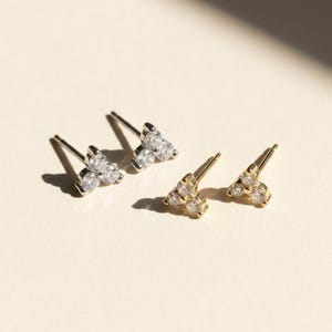 May include: Four pairs of stud earrings. Two pairs are silver-toned, each with three clear, round gemstones arranged in a triangular shape. The other two pairs are gold-toned, also with three clear gemstones in a triangular design.
