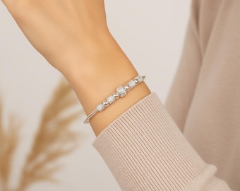 May include: A silver bracelet with a delicate design. The bracelet features a series of small, sparkling beads and a central floral-shaped element. The bracelet is worn on a wrist, showcasing its elegant and refined aesthetic.