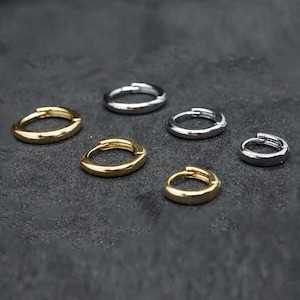 Puede incluir: Seis pendientes de aro pequeños en tonos dorados y plateados están colocados sobre una superficie gris oscuro. Los pendientes son diseños circulares simples, con un cierre de clip. Los pendientes dorados tienen un acabado pulido, mientras que los plateados parecen mate.