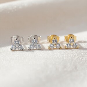 Puede incluir: Dos pares de pendientes de botón. Un par es plateado y el otro dorado. Cada pendiente presenta tres piedras preciosas redondas y transparentes dispuestas en forma triangular. Los pendientes se muestran sobre una superficie blanca texturizada.