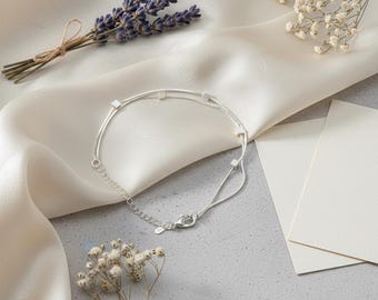 May include: A delicate silver bracelet with small square accents. The bracelet is displayed on a cream-colored satin surface, alongside sprigs of lavender and small white flowers. The bracelet has a lobster clasp closure.