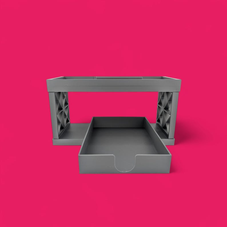 May include: A gray plastic desk organizer with two tiers. The top tier is open and the bottom tier is a drawer. The drawer is pulled out and the organizer is sitting on a pink surface.