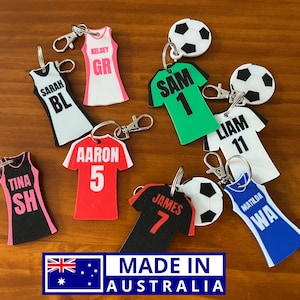 May include: A set of eight colourful keychains featuring sports jerseys with names and numbers. The keychains are made of foam and have a silver metal ring. The keychains are displayed on a wooden surface.