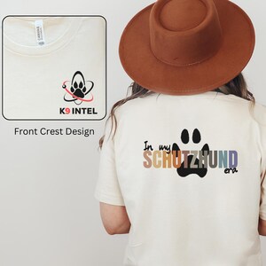 May include: A beige t-shirt with a black paw print and the text "In my Schutzhund era" in rainbow colors on the back. The front of the shirt has a black and red logo with the text "K9 INTEL".