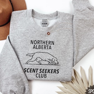Puede incluir: Una sudadera gris claro con el texto "NORTHERN ALBERTA" sobre un dibujo lineal de un perro y "SCENT SEEKERS CLUB" debajo. Un sombrero marrón está en la esquina superior izquierda. La sudadera está etiquetada como "SPORT GREY".