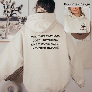 May include: Cream-colored hoodie with the text "AND THERE MY DOG GOES... NEVERING LIKE THEY'VE NEVER NEVERED BEFORE" in black. A small crest with a paw print and the text "K9 INTEL" is on the front. The hoodie has a hood and a kangaroo pocket.