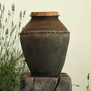 Peut inclure: Un pot en argile brun foncé et patiné, doté d'un corps large et arrondi ainsi que d'une ouverture étroite à bord plat. Le récipient présente une couture horizontale marquée au milieu, avec une texture mate rustique et des tons terreux.