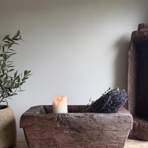 Könnte beinhalten: Eine rechteckige, dunkelbraune, handgeschnitzte Teigschale aus Holz mit einer verwitterten, strukturierten Oberfläche. In der Schale befindet sich eine einzelne weiße Stumpenkerze mit brennender Flamme und ein Bündel getrockneter Lavendel.