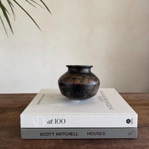 Könnte beinhalten: Eine kleine, dunkelbraune Keramikvase mit rundem Körper und schmalem Hals. Die Vase steht auf zwei Büchern mit den Titeln "AD at 100" und "SCOTT MITCHELL HOUSES". Ein grünes Pflanzenblatt ist oben links zu sehen.