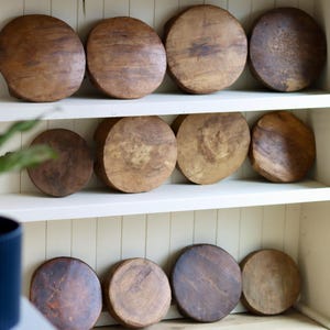 May include: A collection of twelve circular wooden hat blocks arranged on white shelves. Each block features a distinct natural wood grain pattern, varying shades of brown, and a smooth, rounded top surface, showcasing individual textures and wear.