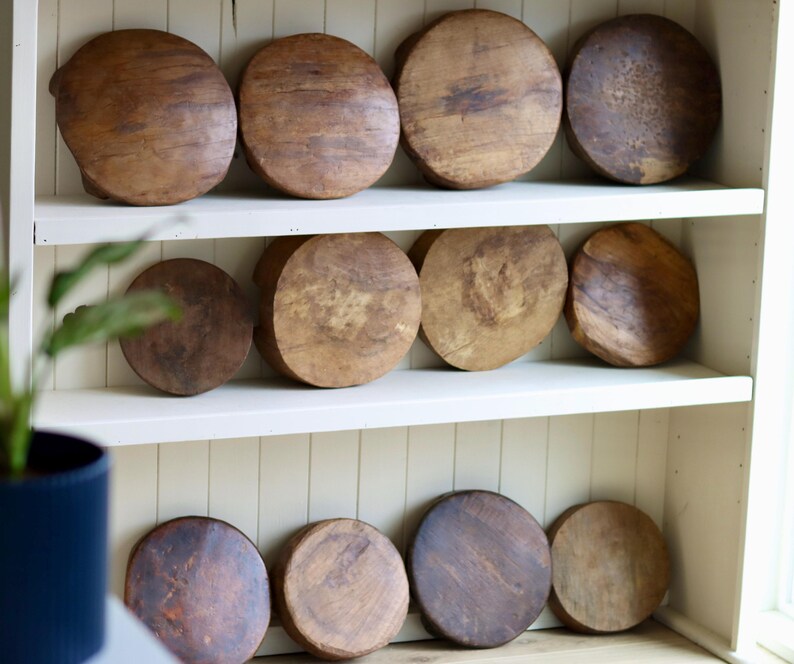 May include: A collection of twelve circular wooden hat blocks arranged on white shelves. Each block features a distinct natural wood grain pattern, varying shades of brown, and a smooth, rounded top surface, showcasing individual textures and wear.