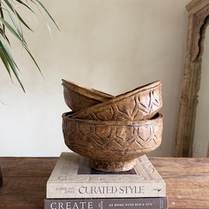 Könnte beinhalten: Drei braune Zierschalen mit geschnitzten geometrischen Mustern sind übereinander gestapelt. Die Schalen stehen auf Büchern mit dem Titel "Curated Style". Die Schalen haben ein rustikales, gealtertes Aussehen.