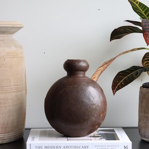 Könnte beinhalten: Eine braune Keramikvase mit rundem Körper und schmalem Hals steht auf einer schwarzen Oberfläche mit einem Buch mit dem Titel "The Modern Apothecary" von Brendan Murdock & Gabriel Weil.