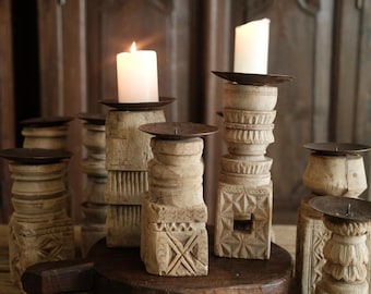 May include: A collection of light-colored, hand-carved wooden candle holders with dark metal plates. Each holder features unique geometric patterns and stacked designs. Two white pillar candles are lit, resting on the wooden bases.