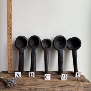 May include: Five dark brown wooden spoons of varying sizes are displayed on a wooden surface. Each spoon has a deep bowl and a long handle. The spoons are labeled A through E. A wooden ruler is on the left side of the image.