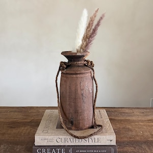 Könnte beinhalten: Eine rustikale, braune Holzvase mit einem Seilgriff, gefüllt mit getrockneten weißen und violett getönten Gräsern. Die Vase steht auf einem Stapel Bücher mit dem Text "CURATED STYLE" auf den Einbänden. Der Gesamteindruck ist vintage und dekorativ.
