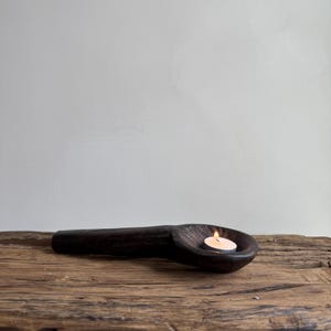May include: A dark brown, wooden candle holder in the shape of a spoon. A small, lit candle sits in the spoon's bowl. The candle holder rests on a rustic, wooden surface, with a plain white background.