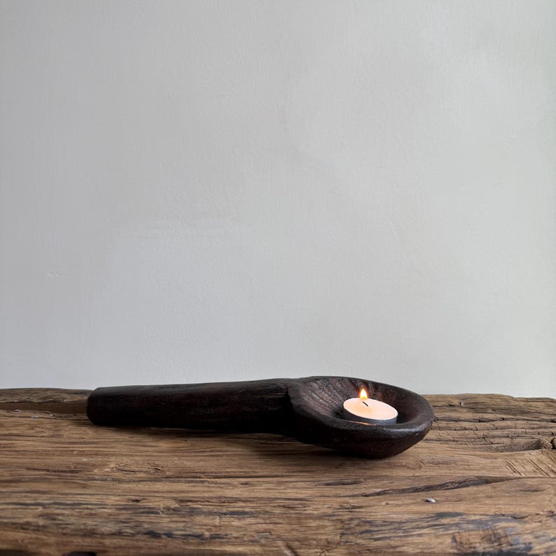 May include: A dark brown, wooden candle holder in the shape of a spoon. A small, lit candle sits in the spoon's bowl. The candle holder rests on a rustic, wooden surface, with a plain white background.