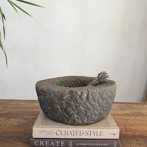 Peut inclure: Un mortier en pierre grise avec une surface texturée, contenant un petit bouquet de lavande séchée. Le mortier repose sur une pile de livres. Les livres ont pour titres "Curated Style" et "Create".
