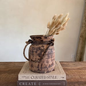 Könnte beinhalten: Eine rustikale Holzvase mit einem Seilgriff und dekorativen Knoten, die getrocknete beige Blumen enthält. Die Vase steht auf zwei Büchern mit den Titeln "CURATED STYLE" und "CREATE". Die Vase hat eine zylindrische Form und ein verwittertes Finish.