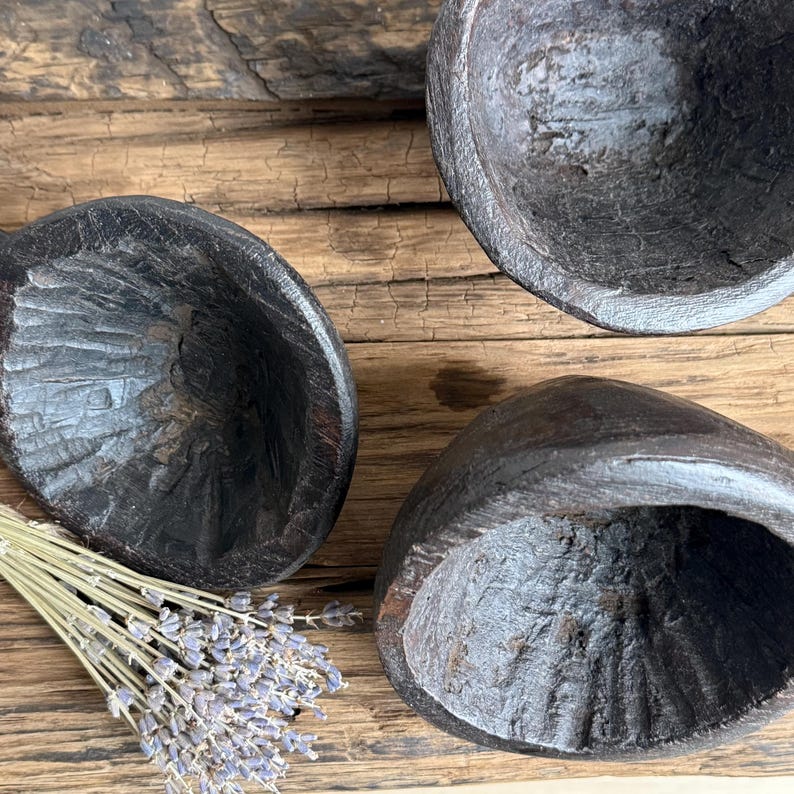 May include: Three dark brown, hand-carved wooden bowls with a textured surface are arranged on a rustic wooden surface. A bundle of dried lavender is placed near one of the bowls. The bowls have a unique, organic shape.