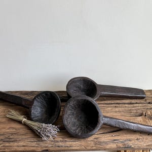 May include: Three dark brown wooden spoons with deep bowls and long handles are arranged on a rustic wooden surface. A bundle of dried lavender is tied with twine and placed near the spoons. The spoons have a carved texture.