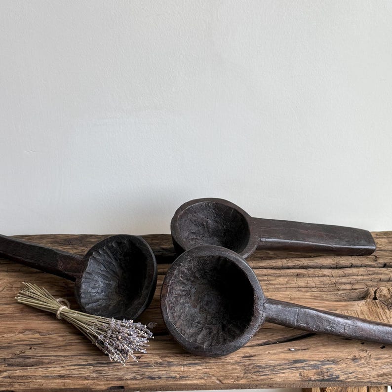 May include: Three dark brown wooden spoons with deep bowls and long handles are arranged on a rustic wooden surface. A bundle of dried lavender is tied with twine and placed near the spoons. The spoons have a carved texture.