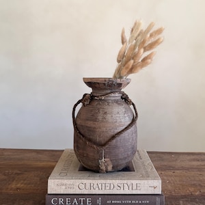 Könnte beinhalten: Eine rustikale, braune Holzvase mit einem Seilgriff, die getrocknete beige Gräser enthält. Die Vase steht auf zwei Büchern mit den Titeln "CURATED STYLE" und "CREATE". Die Vase hat ein verwittertes Aussehen, was ihren Vintage-Charme unterstreicht.