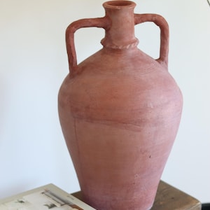 Könnte beinhalten: Eine große, terrakottafarbene Amphorenvase mit zwei Henkeln. Die Vase hat einen abgerundeten Körper und einen schmalen Hals. Sie steht auf einem Holzschemel neben einem Stapel Bücher. Die Vase ist ein dekorativer Gegenstand.