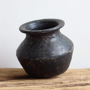 Puede incluir: Un recipiente de cerámica negra, hecho a mano, con un cuerpo redondeado y un borde acampanado. La superficie oscura muestra una textura sutil y patrones decorativos grabados. La olla está sobre una superficie de madera.