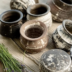 Könnte beinhalten: Eine Sammlung kleiner, runder, rustikaler Keramiktöpfe mit einem abgenutzten Finish in gesprenkelten Weiß- und Dunkelbrauntönen. Jedes Gefäß hat eine weite Öffnung und eine strukturierte Oberfläche mit feinen horizontalen Rillen und Abnutzungsspuren.