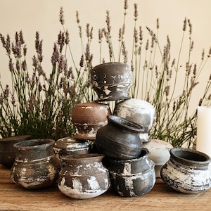 May include: A collection of small, round, dark-toned ceramic pots with a distressed, weathered finish featuring white paint accents. The pots are stacked and arranged in a cluster, displaying varied shades of charcoal, brown, and grey with textured surfaces.